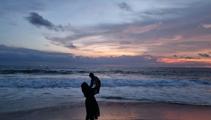 A touching silhouette of a mother lifting her child by the ocean at sunset.