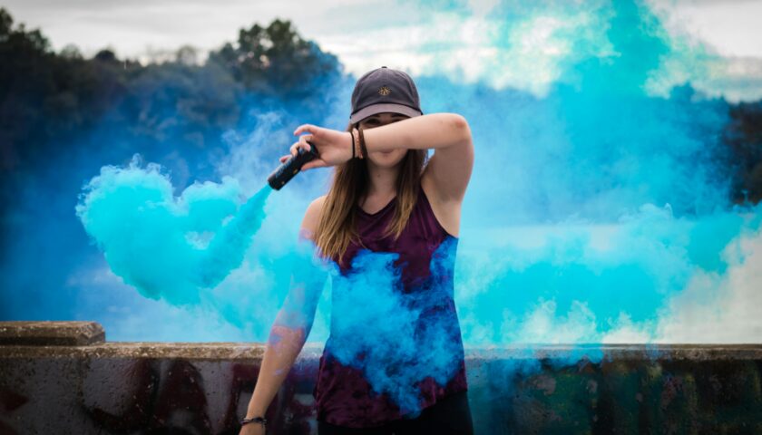 A stylish woman surrounded by bright blue smoke outdoors, creating a dynamic fashion statement.